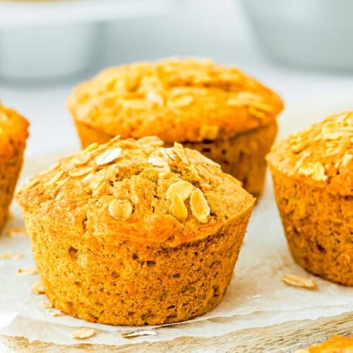 Three golden-brown eggless carrot banana muffins topped with oats rest on a wooden surface.