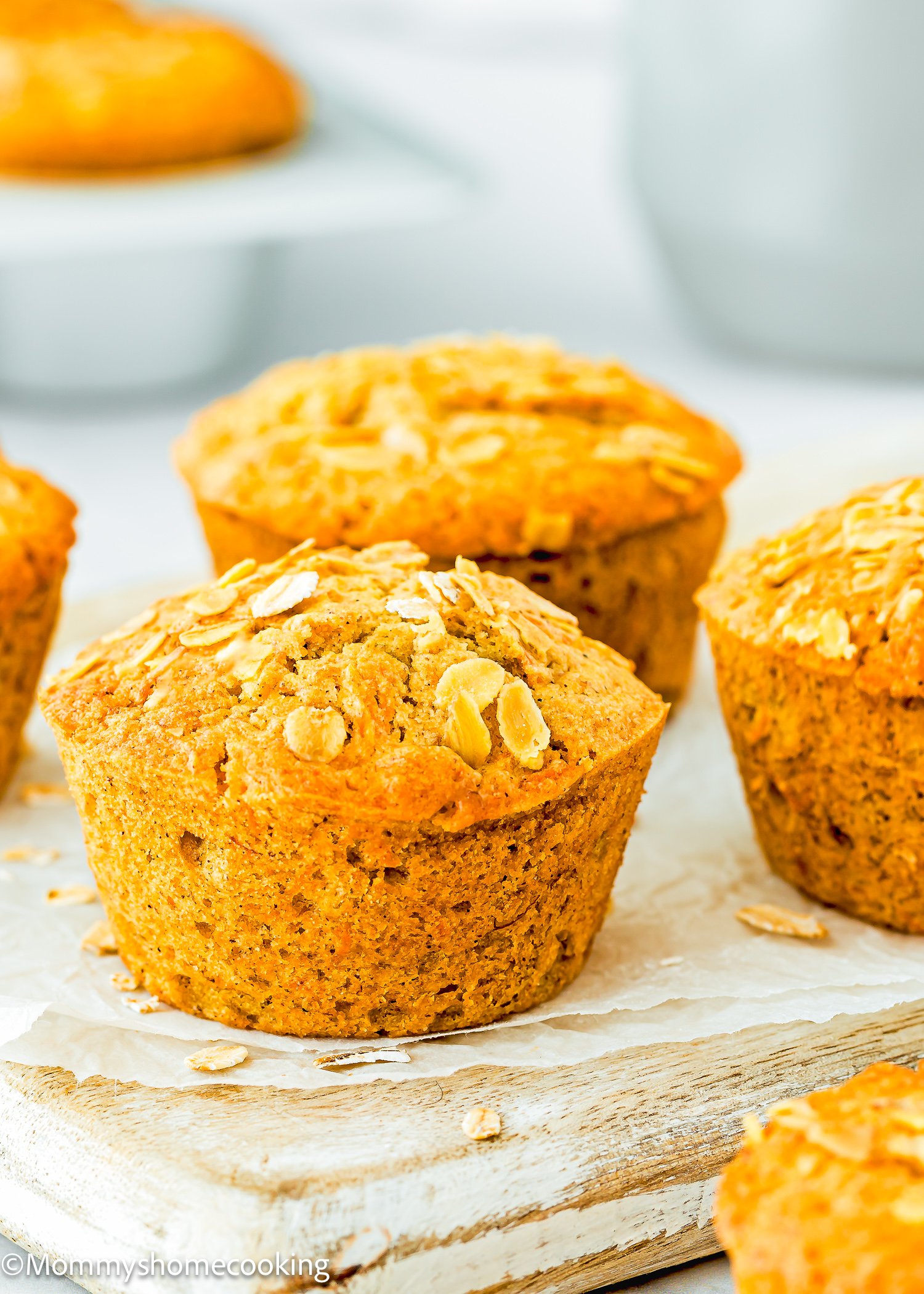 Three golden-brown eggless carrot banana muffins topped with oats rest on a wooden surface.