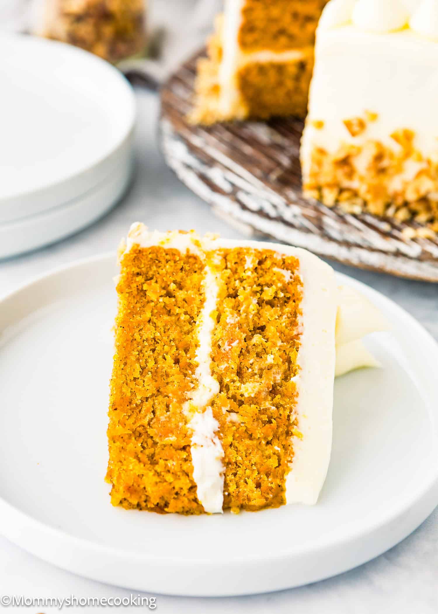 An eggless carrot layer cake sits sliced on a white plate, topped with white frosting. A larger version can be seen in the background.