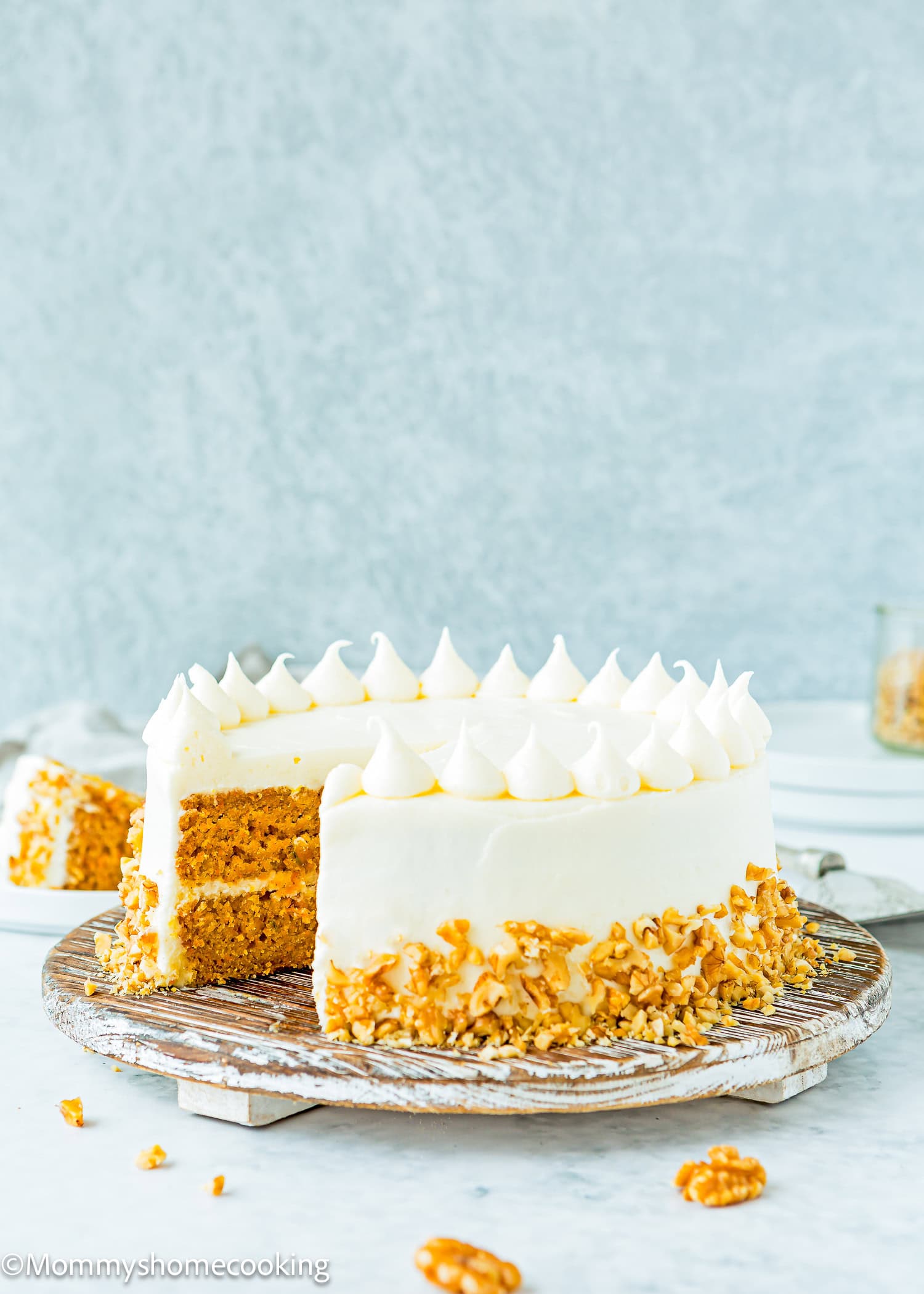 Eggless carrot layer cake with creamy white frosting and crunchy nut garnish, elegantly presented on a wooden stand. A slice is served on a plate in the background.