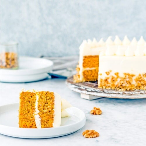 A slice of eggless carrot layer cake with cream cheese frosting sits on a plate, while the whole cake rests on a stand in the background. Walnuts are scattered across the table.