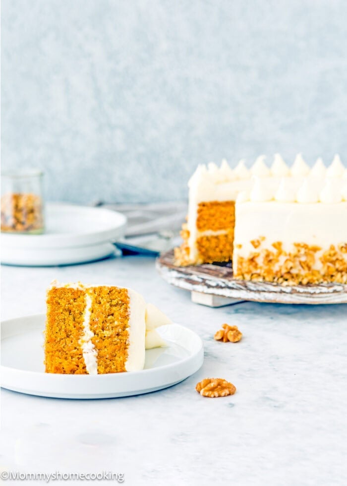 A slice of eggless carrot layer cake with cream cheese frosting sits on a plate, while the whole cake rests on a stand in the background. Walnuts are scattered across the table.