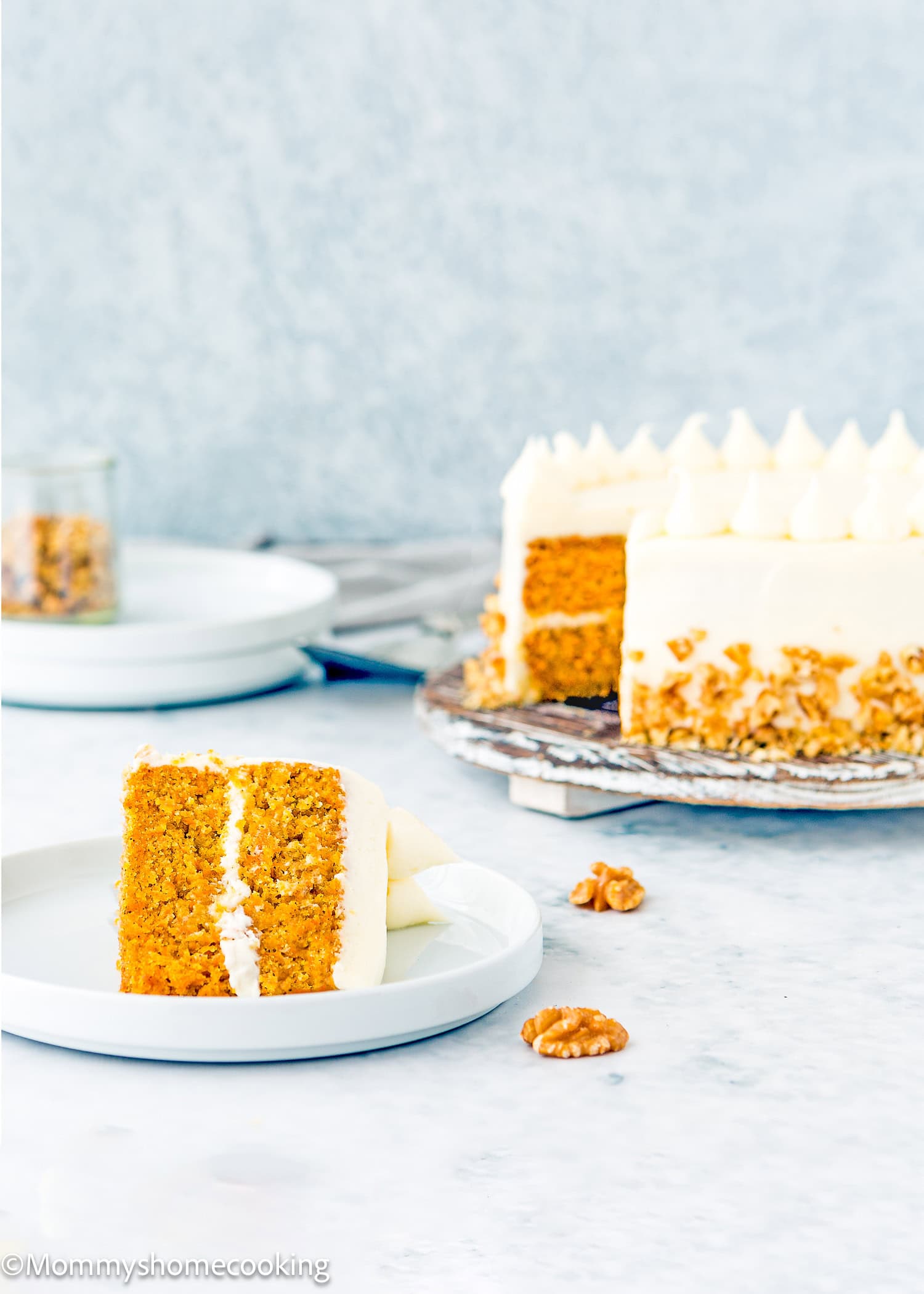 A slice of eggless carrot layer cake with cream cheese frosting sits on a plate, while the whole cake rests on a stand in the background. Walnuts are scattered across the table.