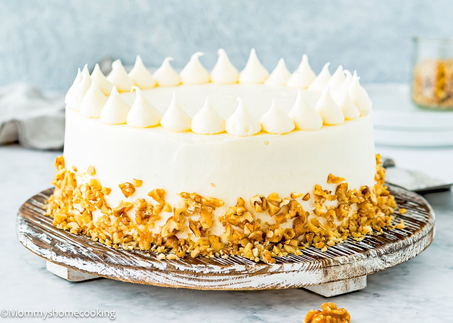 An eggless carrot layer cake with white frosting is topped with whipped cream dollops. Chopped walnuts are pressed around the base, adding a delightful crunch. The cake elegantly sits on a wooden stand placed on a marble surface.