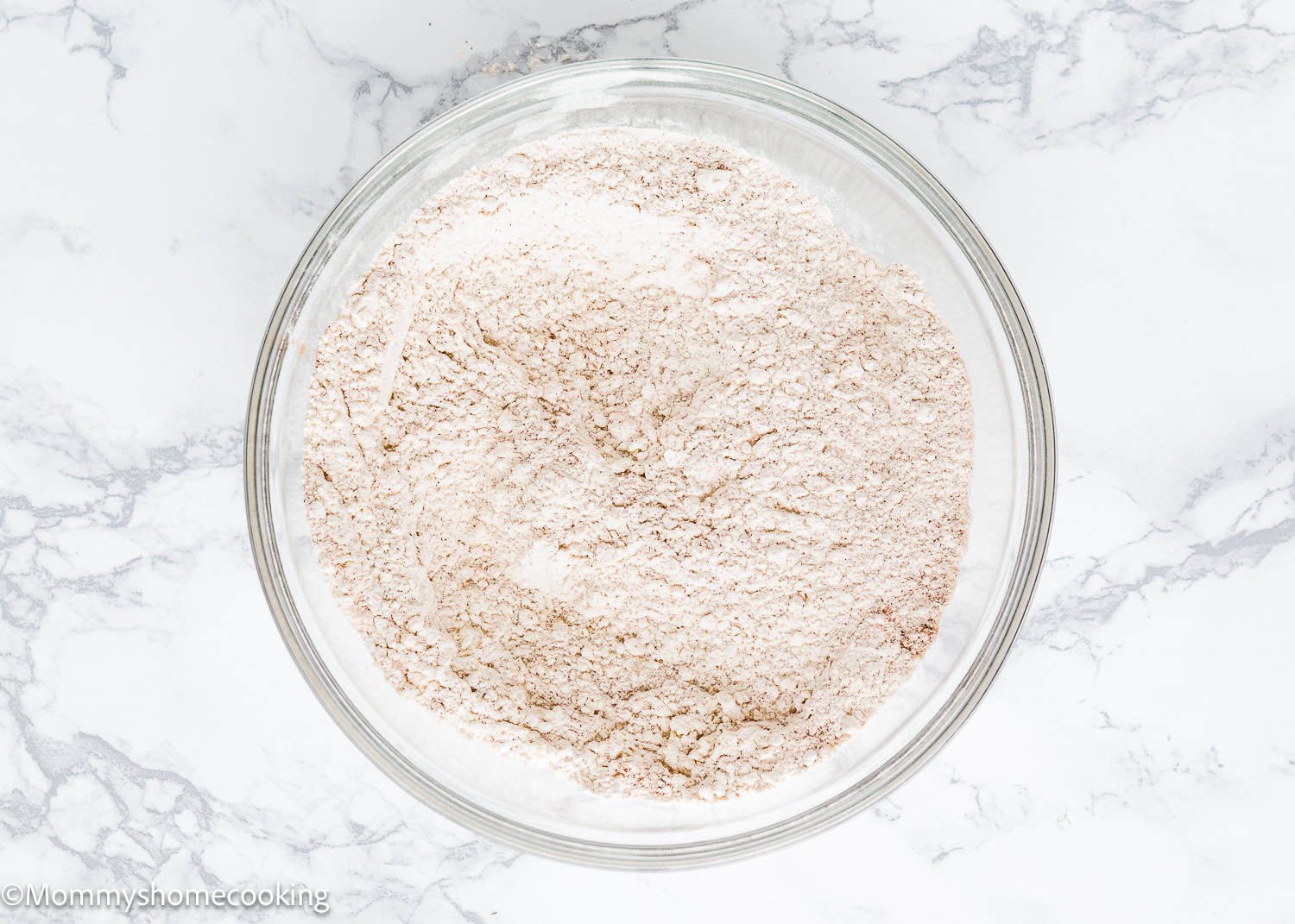 A glass bowl filled with dry ingredients sits on a marble countertop, ready to be transformed into an eggless carrot layer cake.