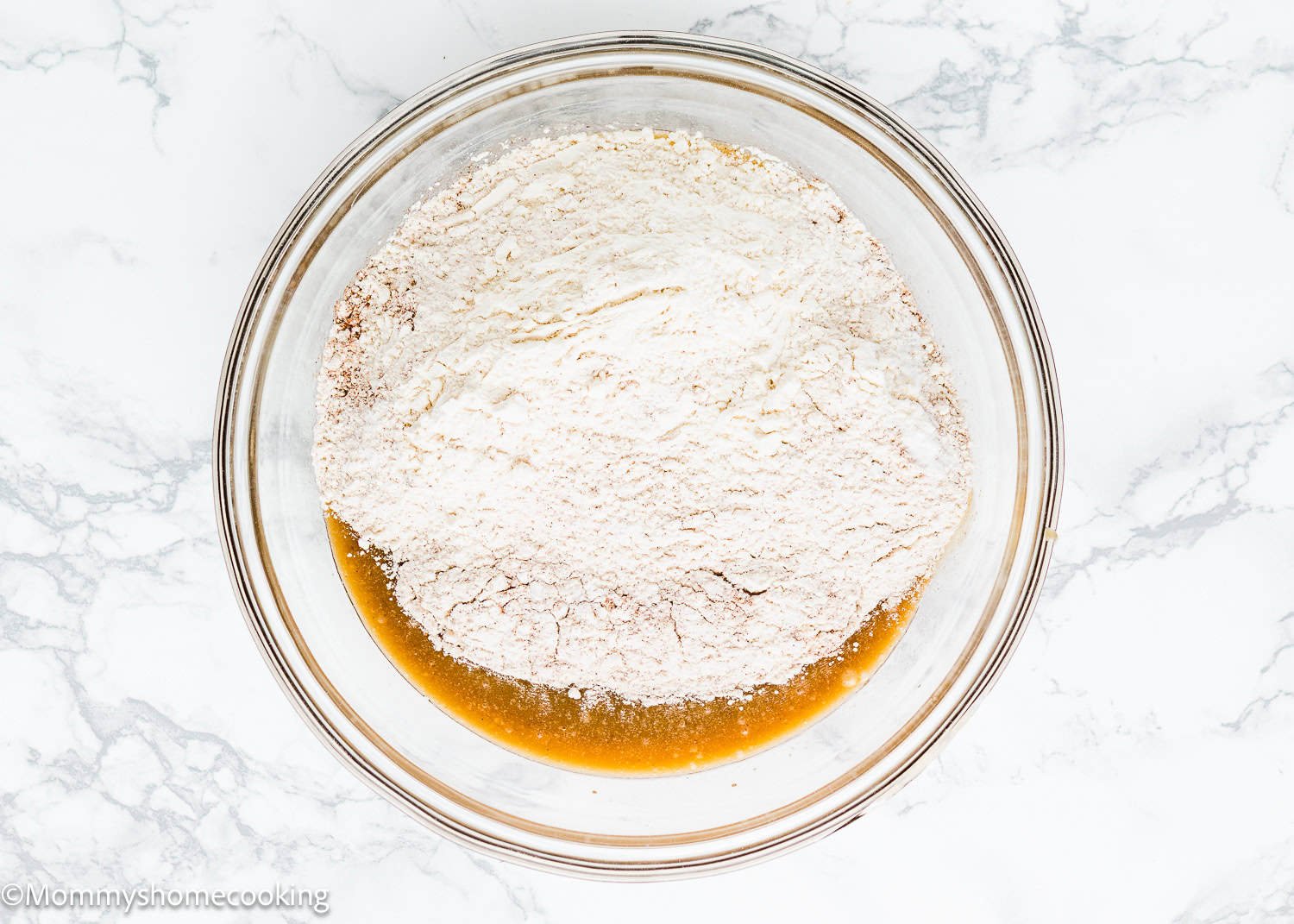 A glass bowl on a marble surface holds a mixture dry and wet ingredients, the first step in creating an eggless carrot layer cake.