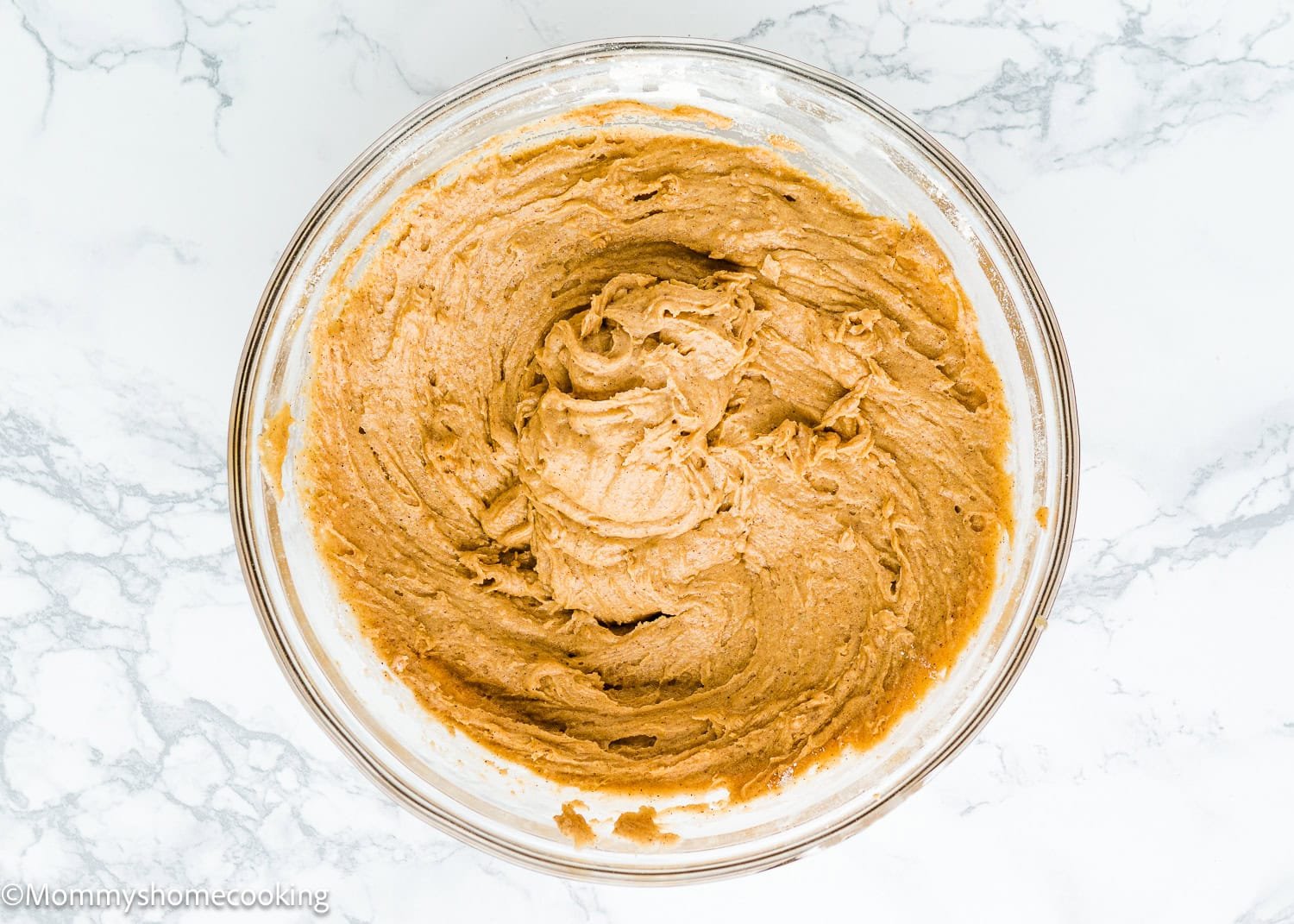A glass bowl filled with eggless carrot batter sits on a marble surface, ready to create a delightful carrot layer cake.
