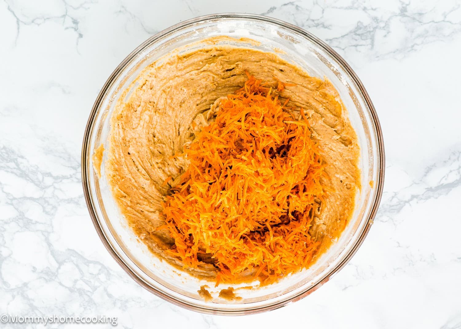 A glass bowl with batter for an eggless carrot layer cake is topped with shredded carrots, resting on a light marble surface.
