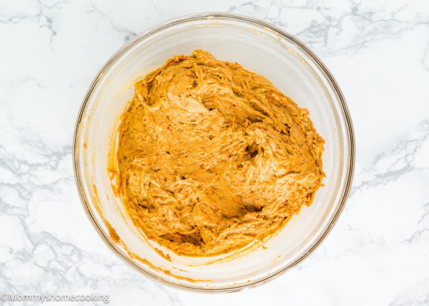 A glass bowl filled with thick brown batter for an eggless carrot layer cake sits on a marble surface.