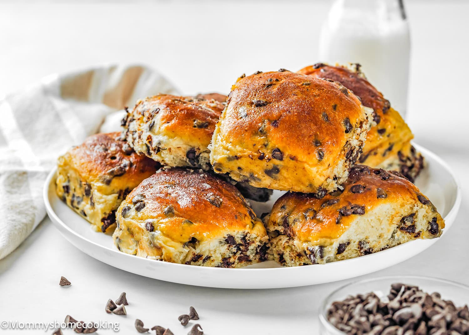 A plate of golden-brown Eggless Chocolate Chip Brioche Rolls is stacked on a white surface, with a glass bottle of milk and a striped cloth in the background.