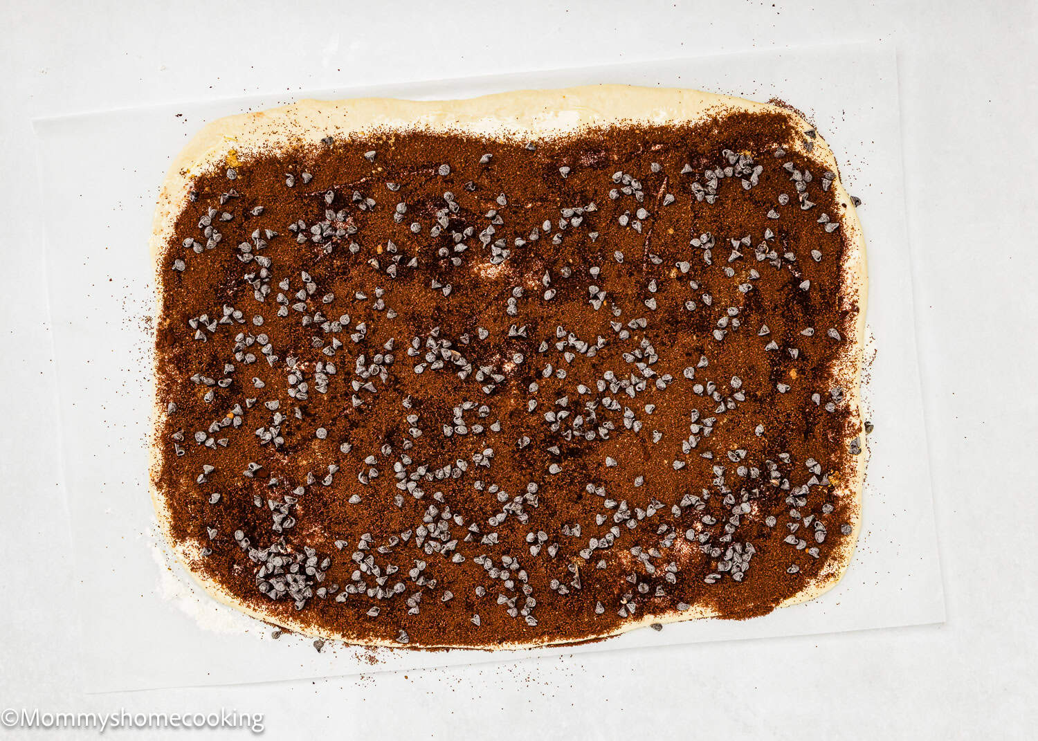 A rectangular sheet of dough for Eggless Chocolate Cinnamon Rolls topped with brown sugar, cinnamon, and scattered mini chocolate chips on a white surface.