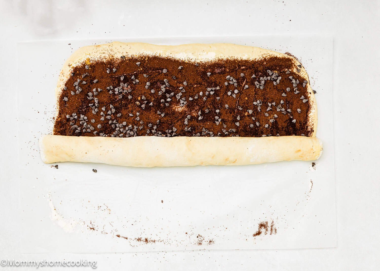 A sheet of dough, partially rolled up, with a filling of cinnamon, sugar, and chocolate chips on a white surface—perfect for making Eggless Chocolate Cinnamon Rolls.