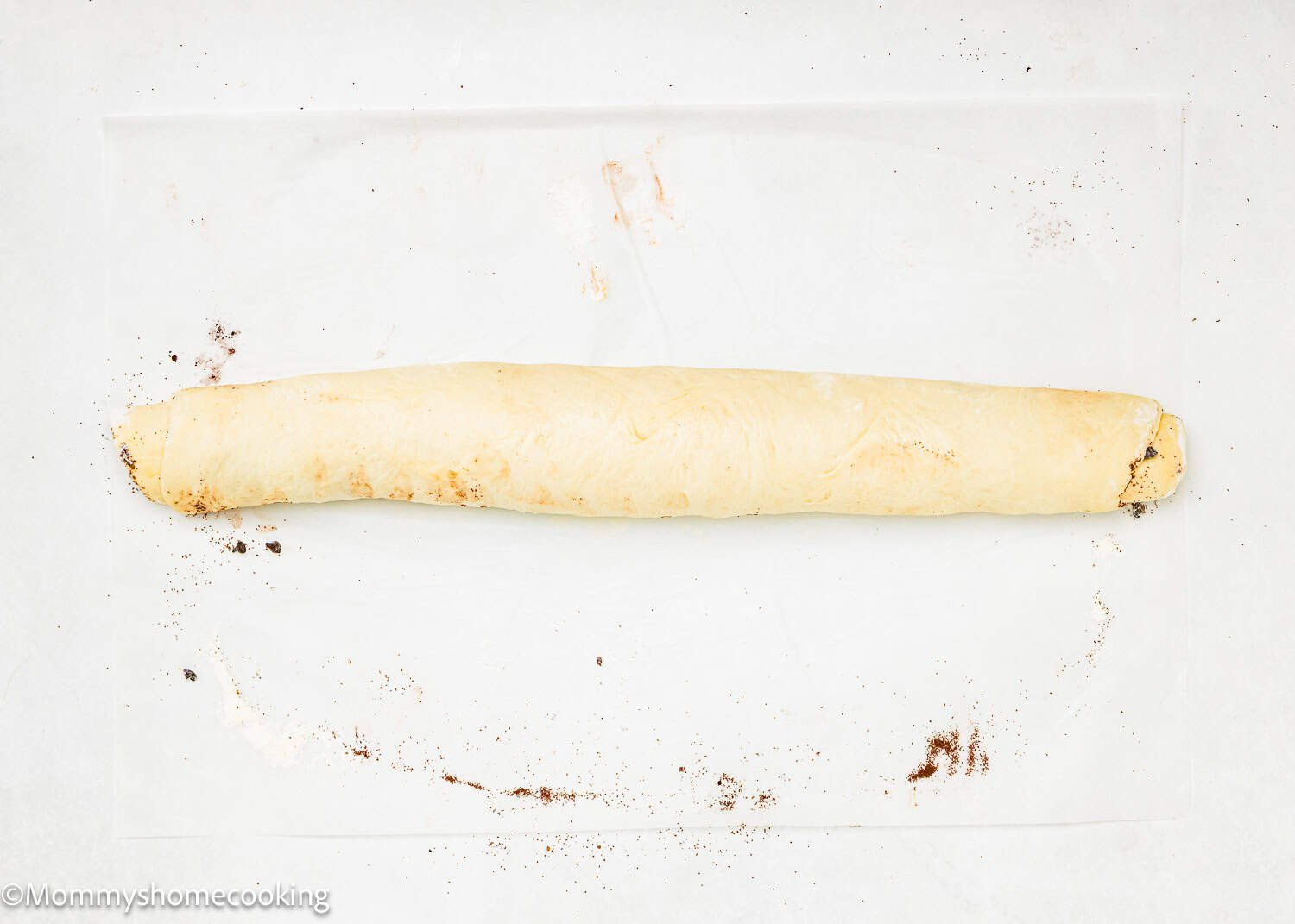 A rolled log of dough with some cinnamon and sugar residue on parchment paper, perfect for making Eggless Chocolate Cinnamon Rolls before slicing or baking.