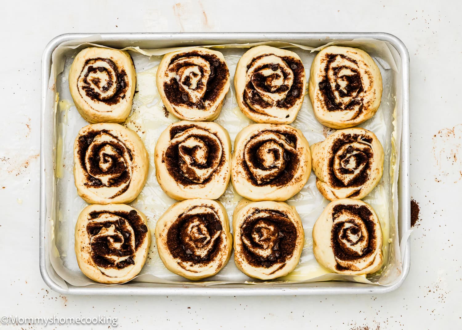 A metal baking sheet lined with parchment paper holds twelve unbaked Eggless Chocolate Cinnamon Rolls arranged in three rows.