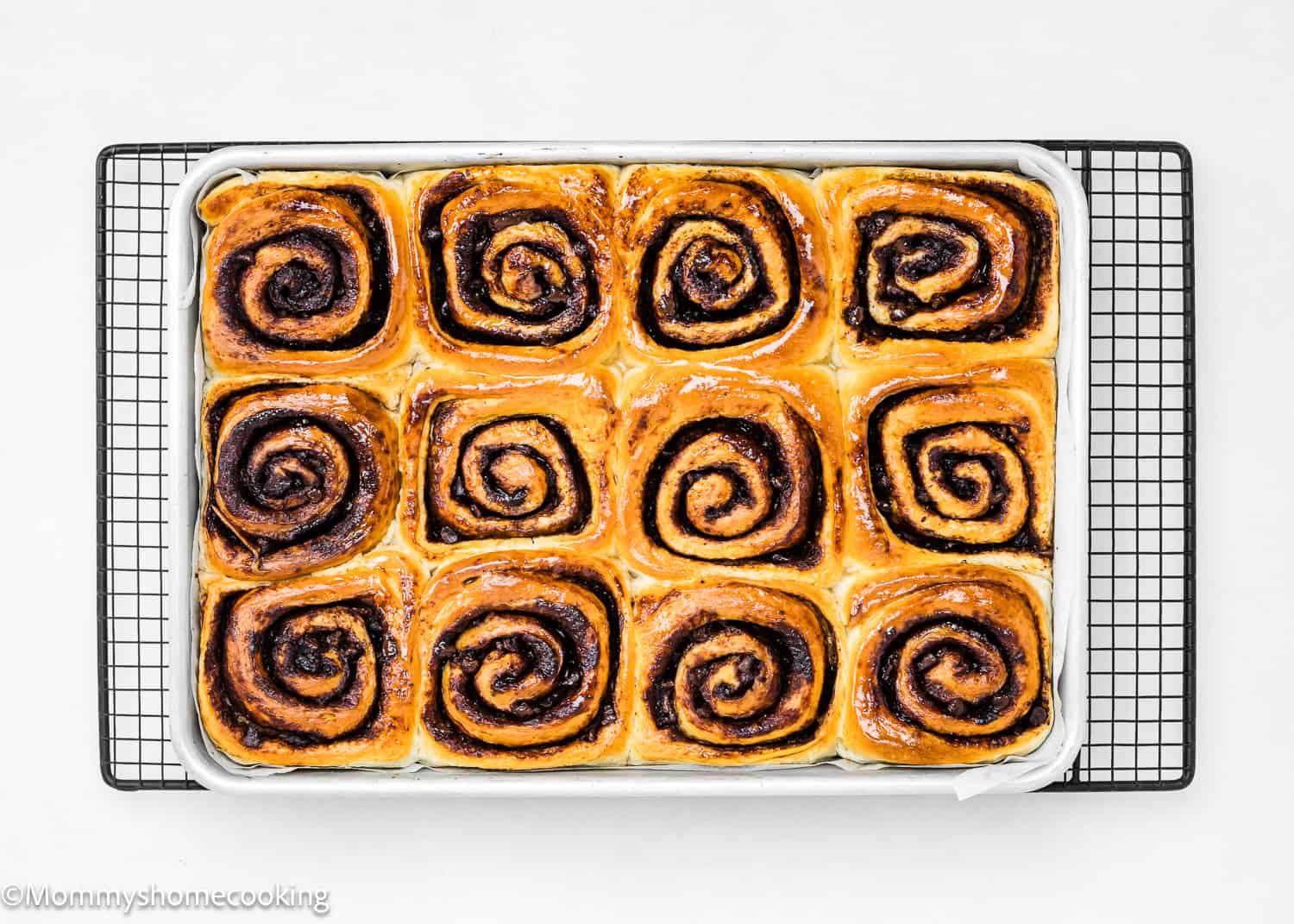A tray of twelve freshly baked Eggless Chocolate Cinnamon Rolls with a golden brown, shiny glaze rests on a cooling rack against a white background.