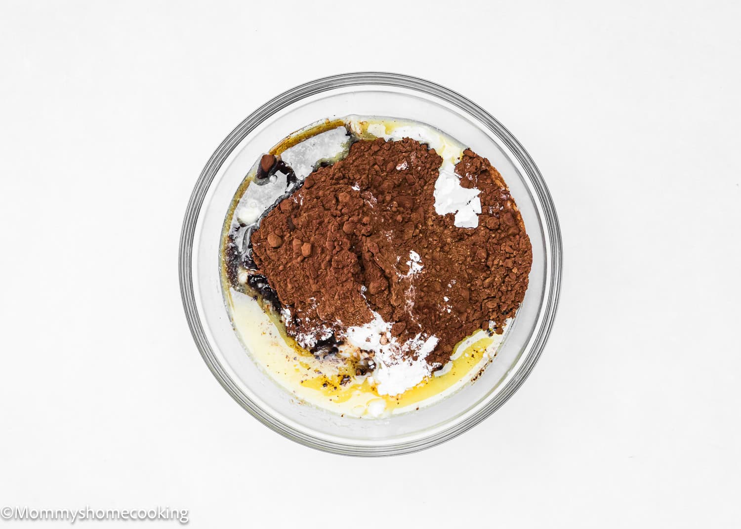 A glass bowl containing cocoa powder, melted butter, sugar, and other baking ingredients for Eggless Chocolate Sweet Rolls on a white background.