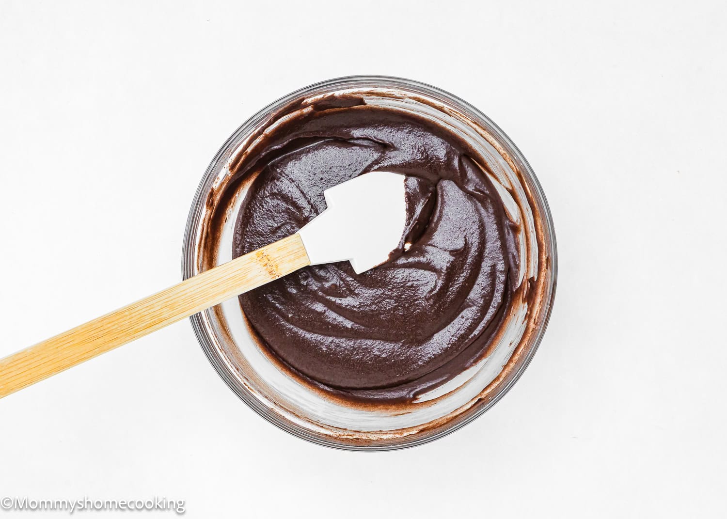 A bowl of thick chocolate batter for Eggless Chocolate Sweet Rolls with a spatula resting on the rim, viewed from above on a white background.