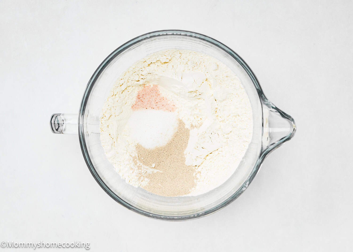A glass mixing bowl containing flour, yeast, sugar, and salt on a white surface, ready to make Eggless Chocolate Cinnamon Rolls.