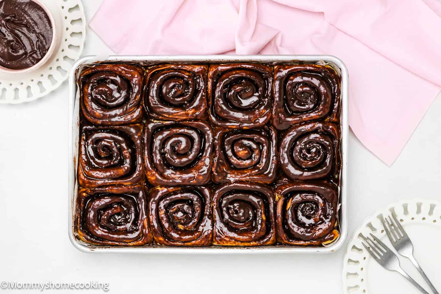 A rectangular baking pan with 12 Eggless Chocolate Sweet Rolls, two plates, a cup of chocolate sauce, forks, and a pink cloth nearby.
