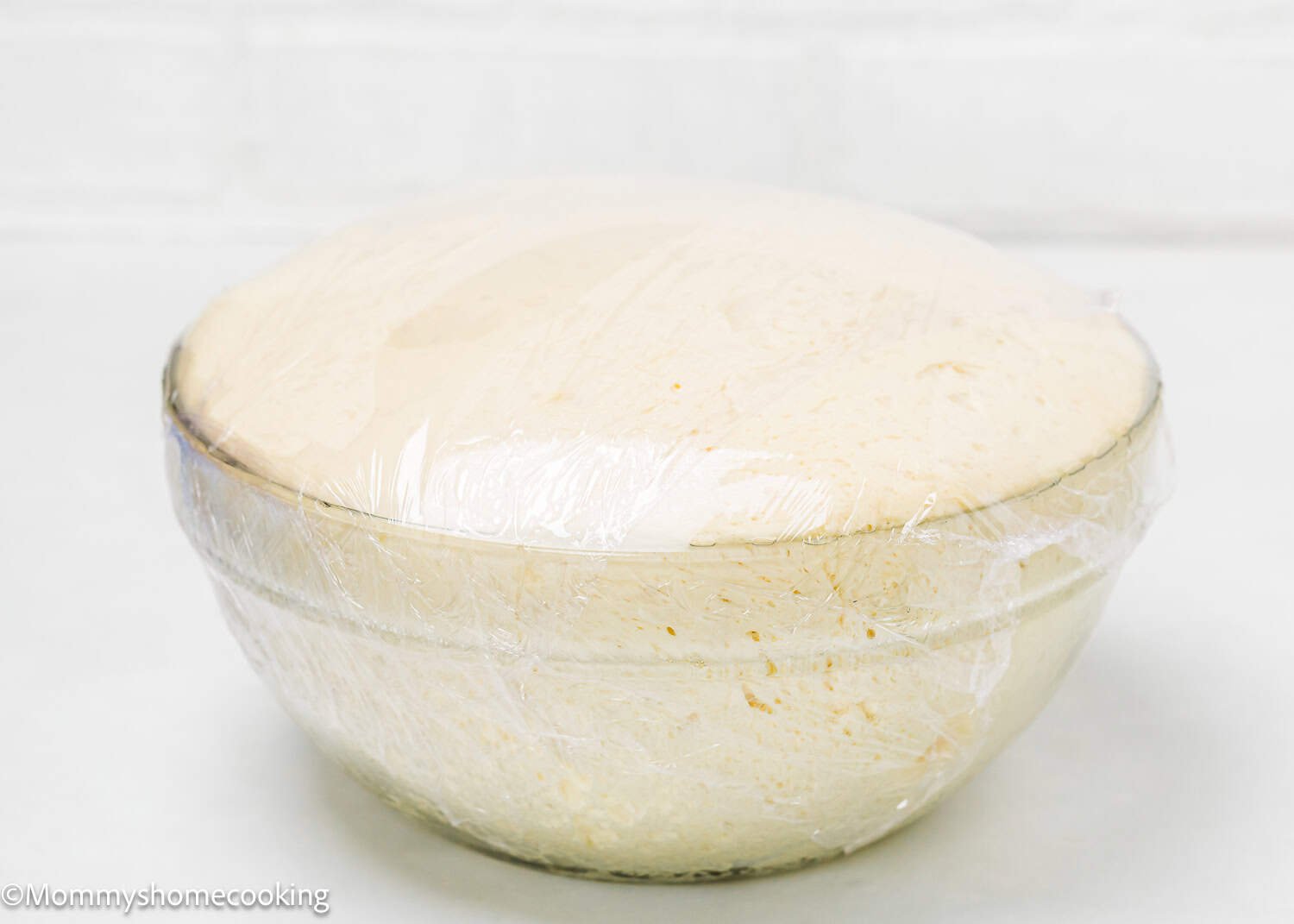 A glass bowl filled with risen dough for Eggless Chocolate Cinnamon Rolls, covered with plastic wrap, sits on a white surface.