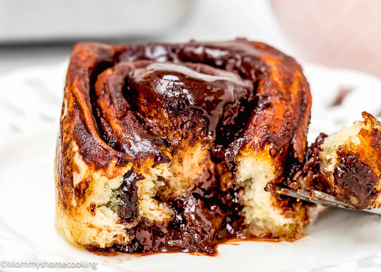 A close-up of Eggless Chocolate Cinnamon Rolls on a white plate, with a fork cutting into the roll and melted chocolate filling visible.