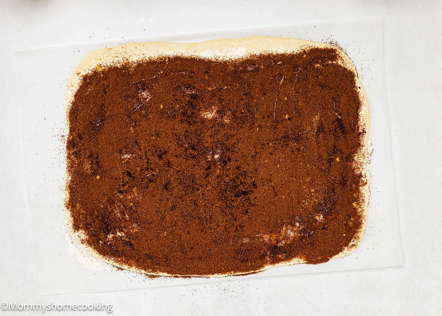 Rectangular sheet of dough spread with a cinnamon and sugar mixture on a parchment-lined surface, perfect for making Eggless Chocolate Cinnamon Rolls.