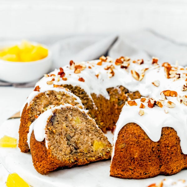 A sliced Eggless Hummingbird Bundt Cake with white icing and chopped pecans sits on a white surface, with pineapple pieces and a bowl of pineapple in the background.
