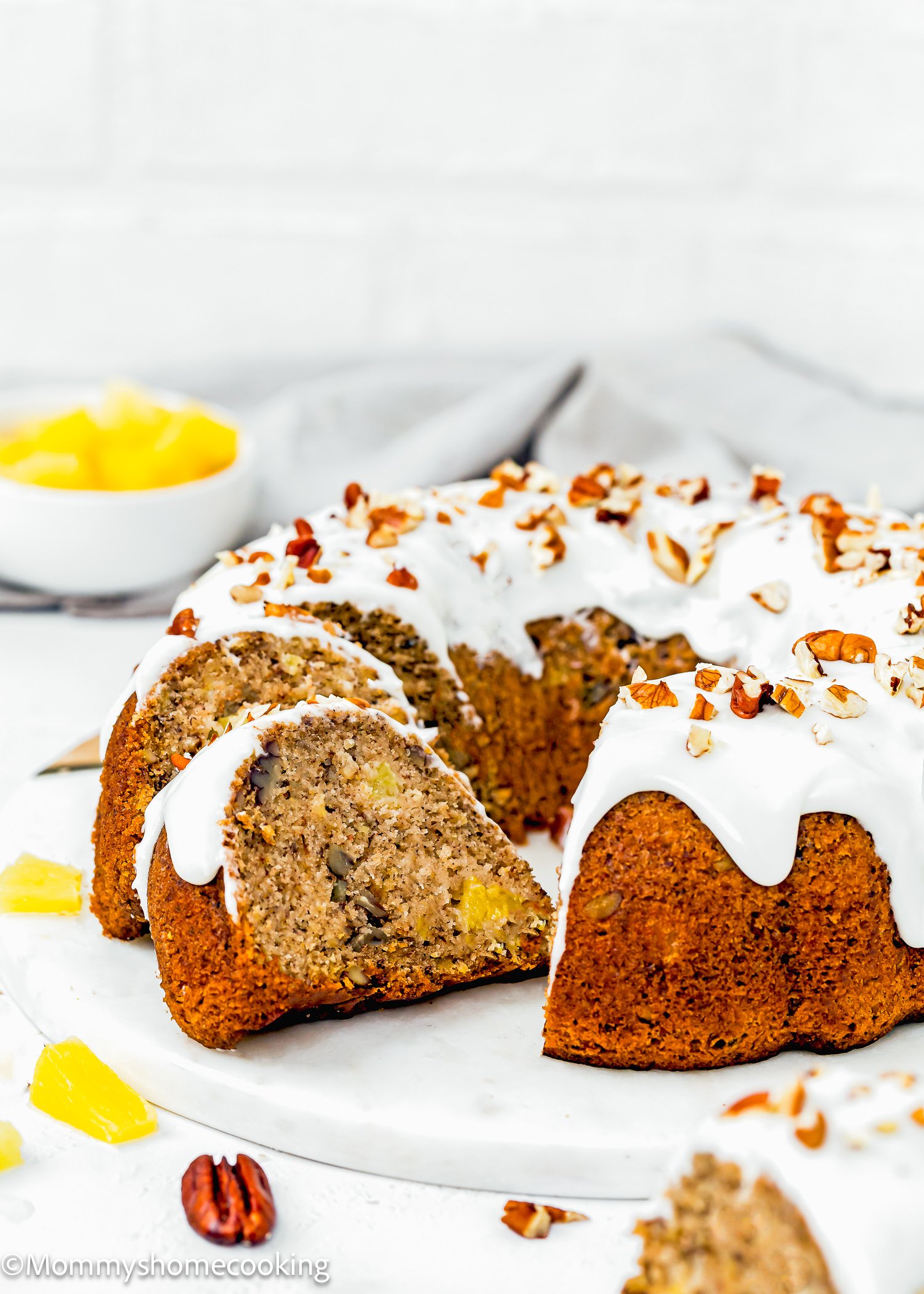 A sliced Eggless Hummingbird Bundt Cake with white icing and chopped pecans sits on a white surface, with pineapple pieces and a bowl of pineapple in the background.