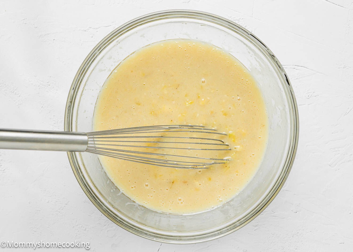 A glass bowl containing a wet ingredients to make Eggless Hummingbird Bundt Cake being whisked with a metal whisk on a white surface.