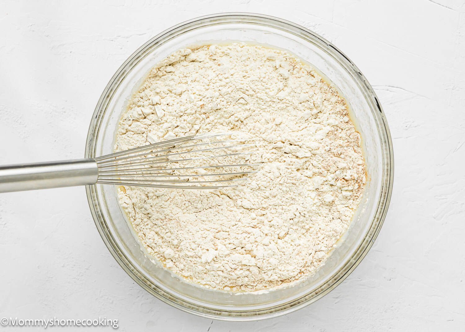 A metal whisk rests in a glass bowl filled with dry and wet ingredients, ready to make an Eggless Hummingbird Bundt Cake.