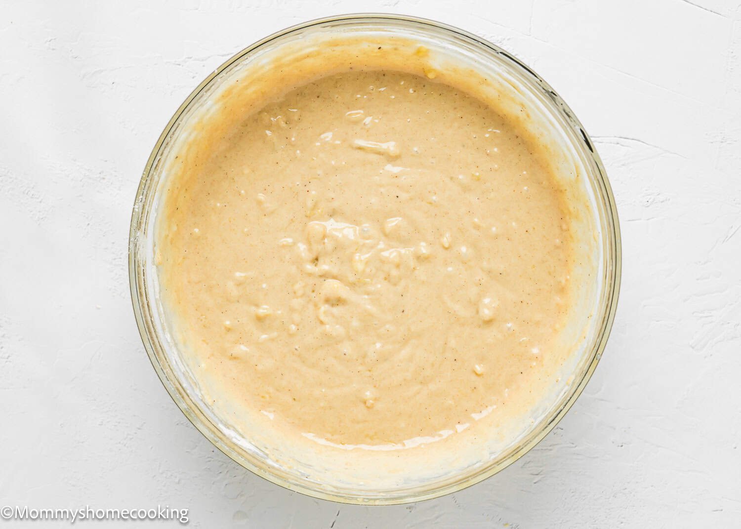 A glass bowl filled with thick, light brown Eggless Hummingbird Bundt Cake batter sits on a white surface.