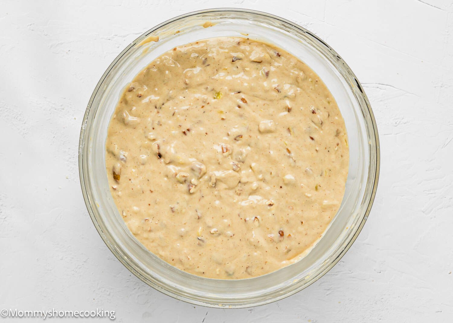 A glass bowl filled with thick, beige Eggless Hummingbird Bundt Cake batter containing visible chunks, placed on a white surface.