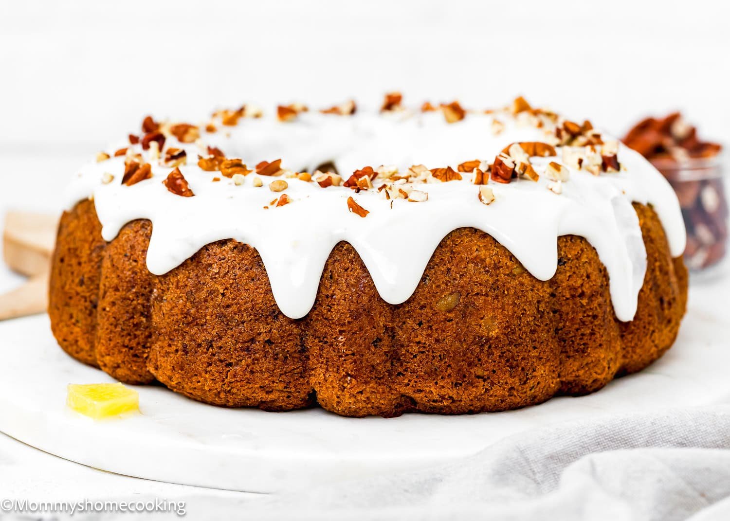 An Eggless Hummingbird Bundt Cake topped with white icing and chopped nuts sits on a white surface; a small cube of butter is nearby.