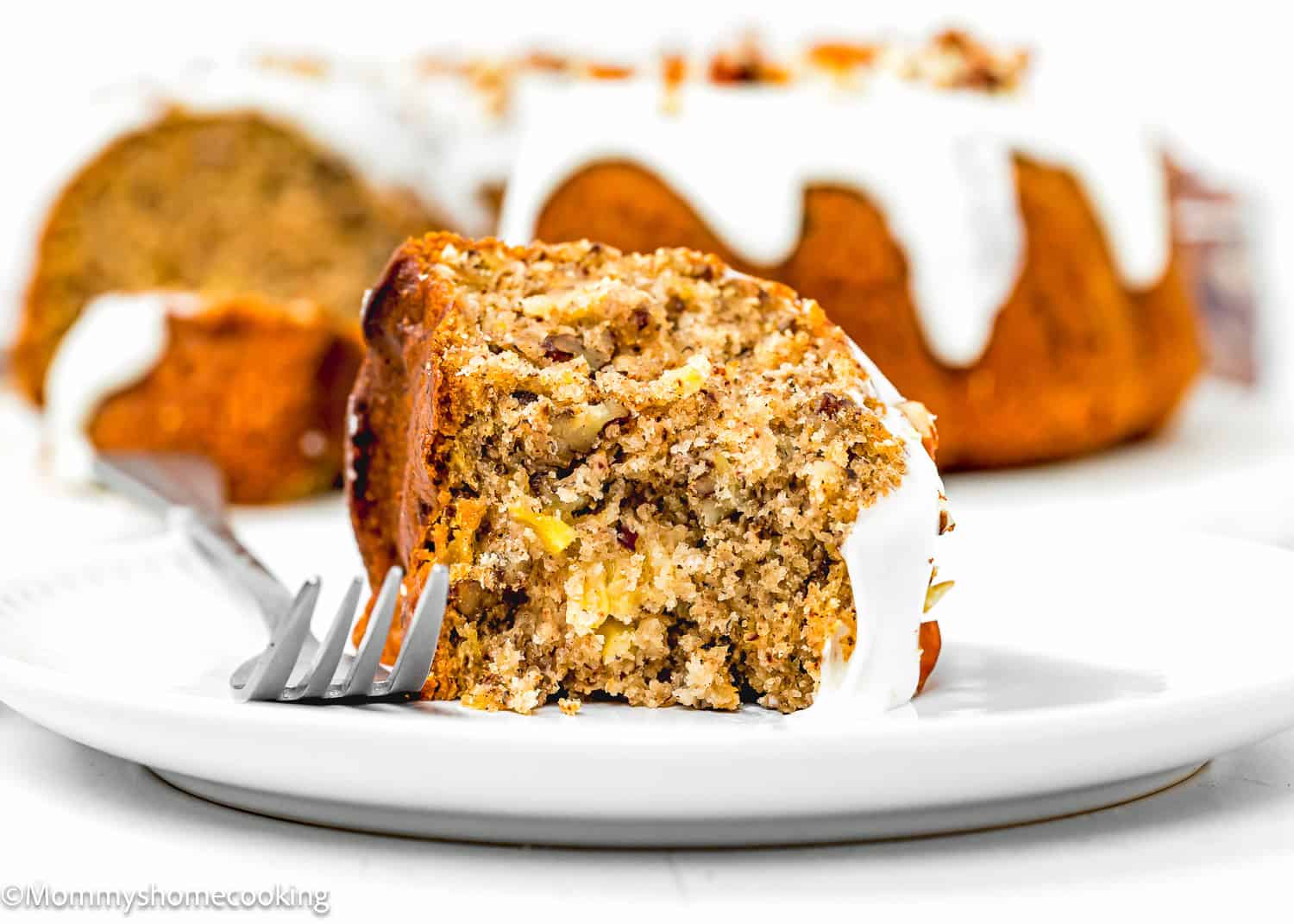 A slice of Eggless Hummingbird Bundt Cake with visible apple pieces sits on a white plate, a fork beside it and the rest of the cake in the background.