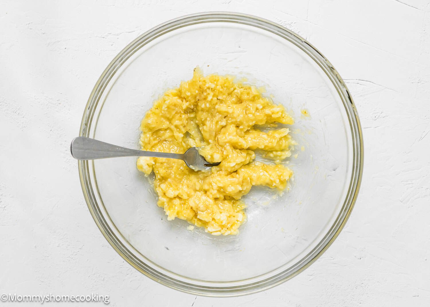 A glass bowl containing mashed bananas with a fork on a white surface.