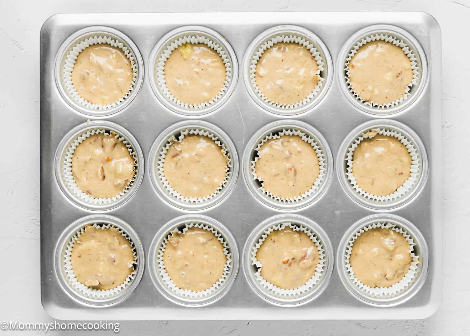 A muffin tin filled with twelve paper liners, each holding unbaked Eggless Hummingbird Cupcakes batter with visible nut pieces.