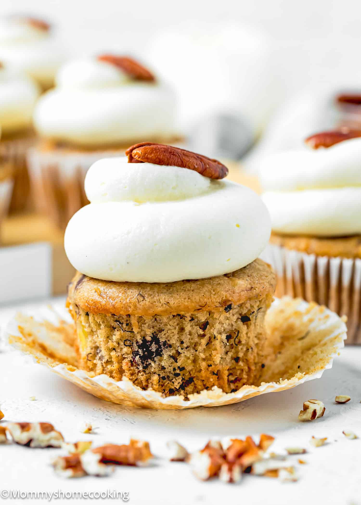 An Eggless Hummingbird Cupcake with white frosting and a pecan on top, partially unwrapped from its paper liner, with chopped pecans scattered nearby.