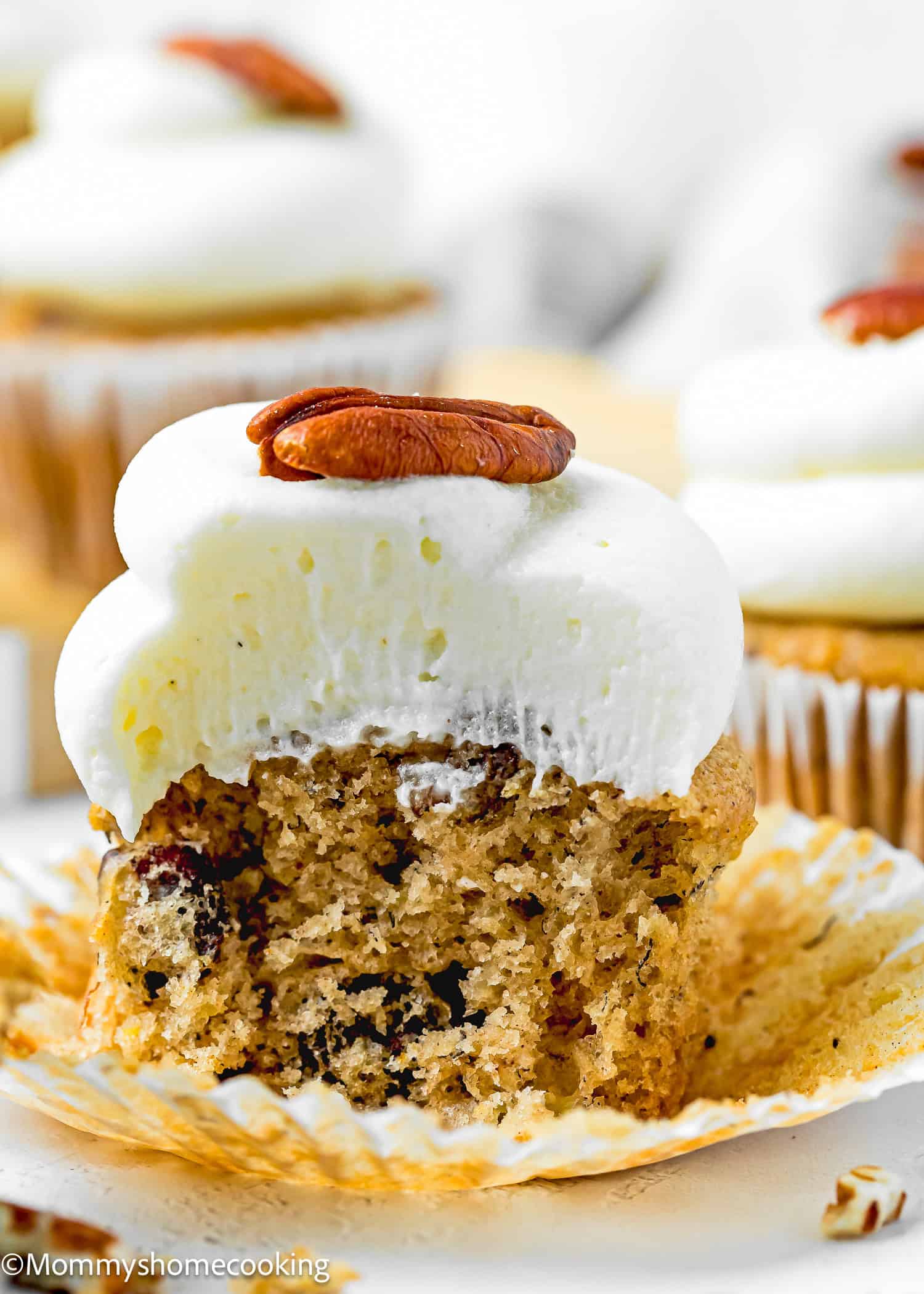 An Eggless Hummingbird Cupcake with white frosting and a pecan on top, partially unwrapped and bitten to reveal a moist interior with visible pecan pieces.
