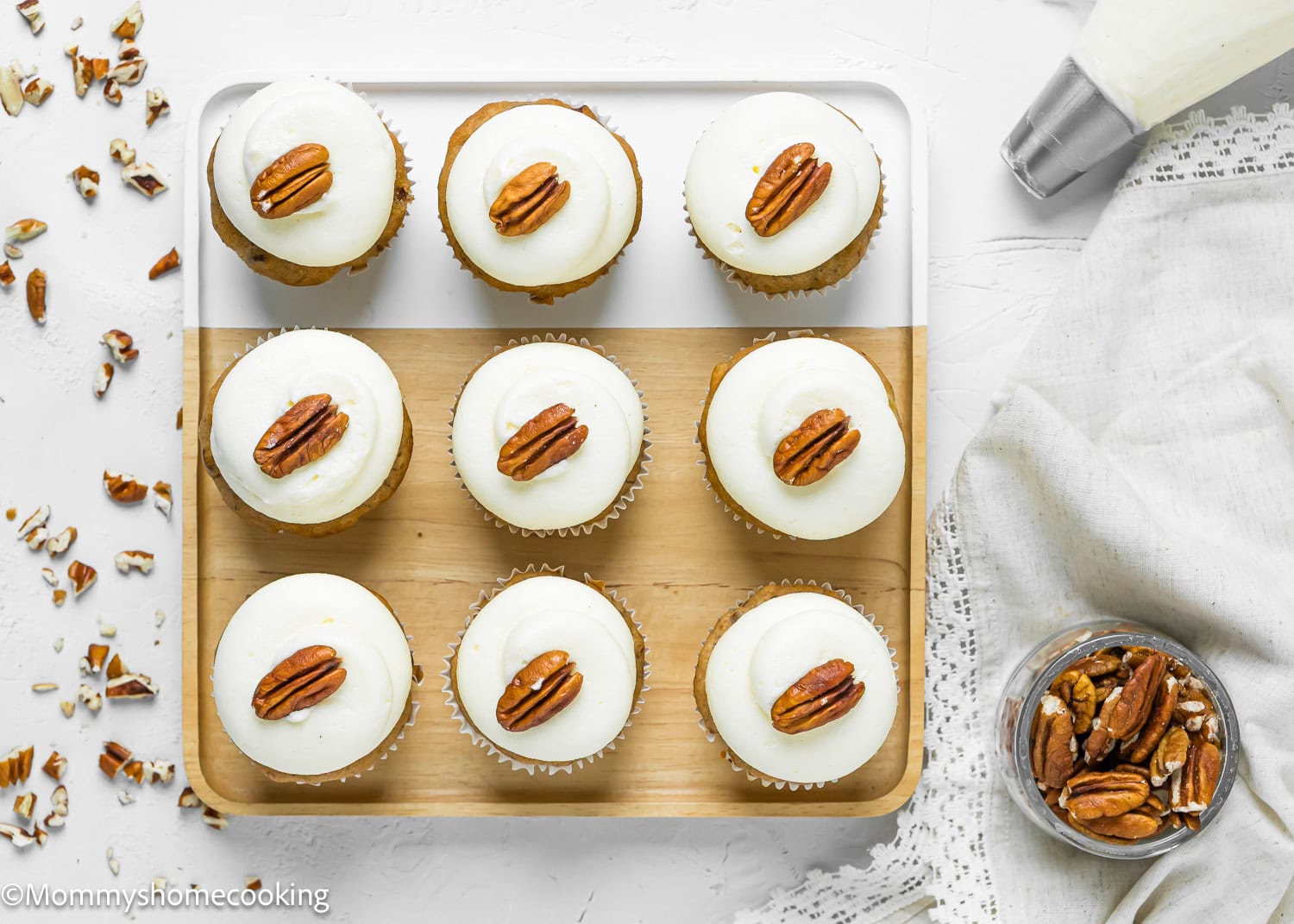 Nine Eggless Hummingbird Cupcakes with white frosting and a single pecan on top are arranged on a wooden tray, with a piping bag, napkin, and a jar of pecans nearby.