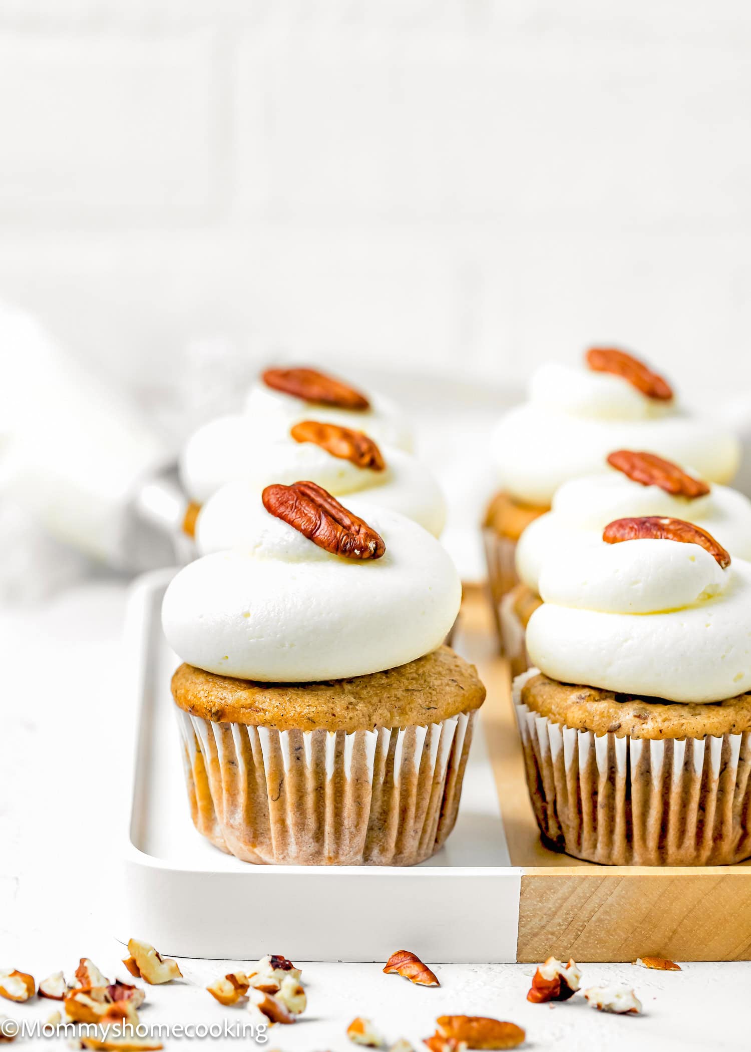 Four Eggless Hummingbird Cupcakes topped with white frosting and a pecan half are arranged on a white tray, with chopped pecans scattered nearby.