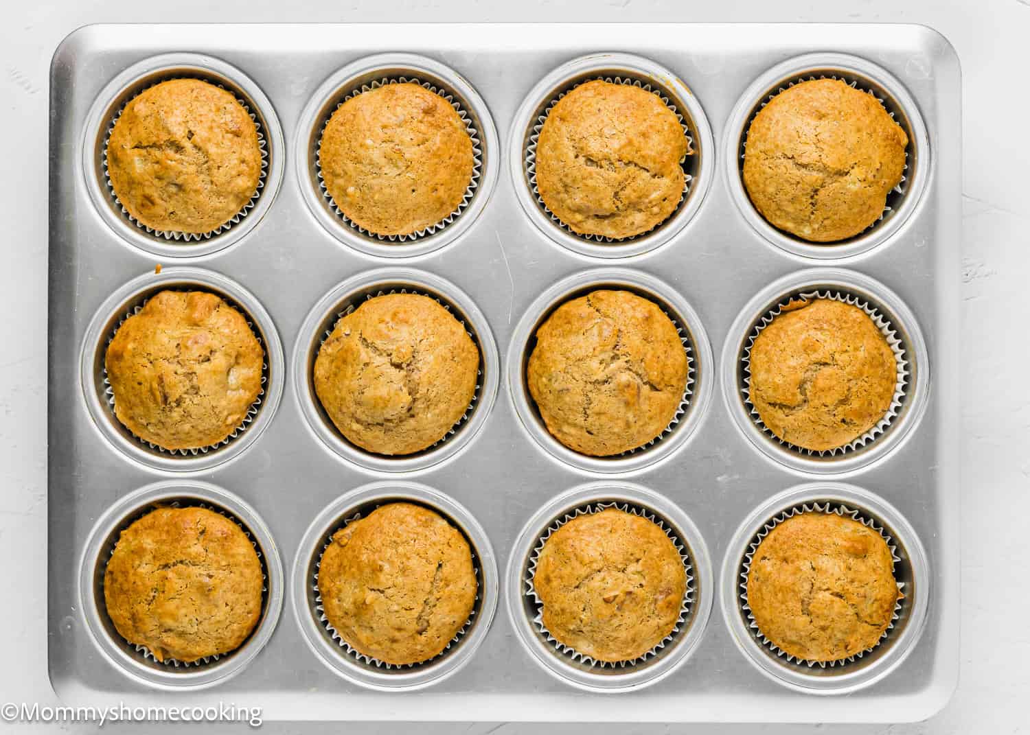 A metal muffin tray holding twelve evenly baked Eggless Hummingbird Cupcakes in paper liners, viewed from above on a light surface.