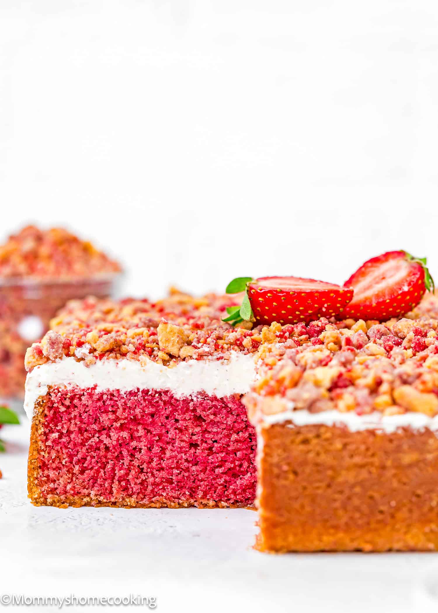 An Eggless Strawberry Crunch Cake, rectangular with a layer of white frosting and crumb topping, is garnished with halved strawberries. A slice is removed to reveal its soft pink interior.