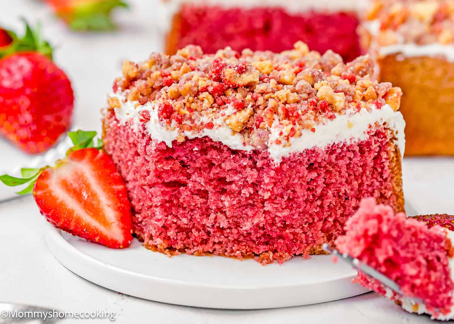 A slice of Eggless Strawberry Crunch Cake with pink sponge, white frosting, and crumb topping sits on a white plate with a halved strawberry beside it.