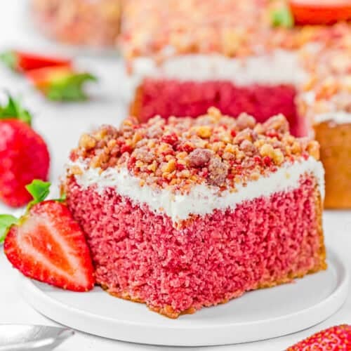 A slice of Eggless Strawberry Crunch Cake with white frosting and crumb topping sits on a white plate, garnished with a fresh strawberry. Other cake slices and strawberries are in the background.