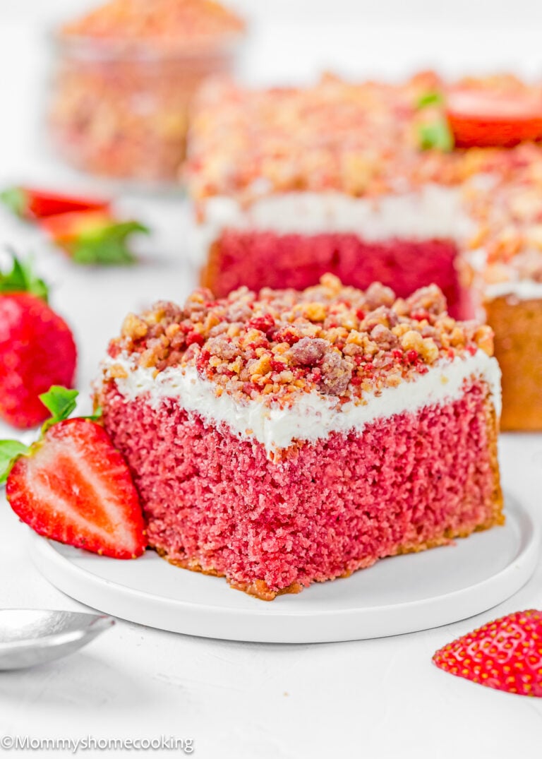 A slice of Eggless Strawberry Crunch Cake with white frosting and crumb topping sits on a white plate, garnished with a fresh strawberry. Other cake slices and strawberries are in the background.