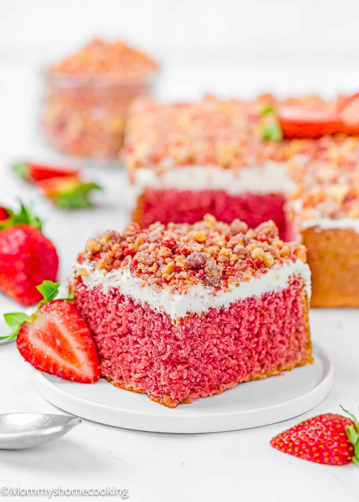A slice of Eggless Strawberry Crunch Cake with white frosting and crumb topping sits on a white plate, garnished with a fresh strawberry. Other cake slices and strawberries are in the background.