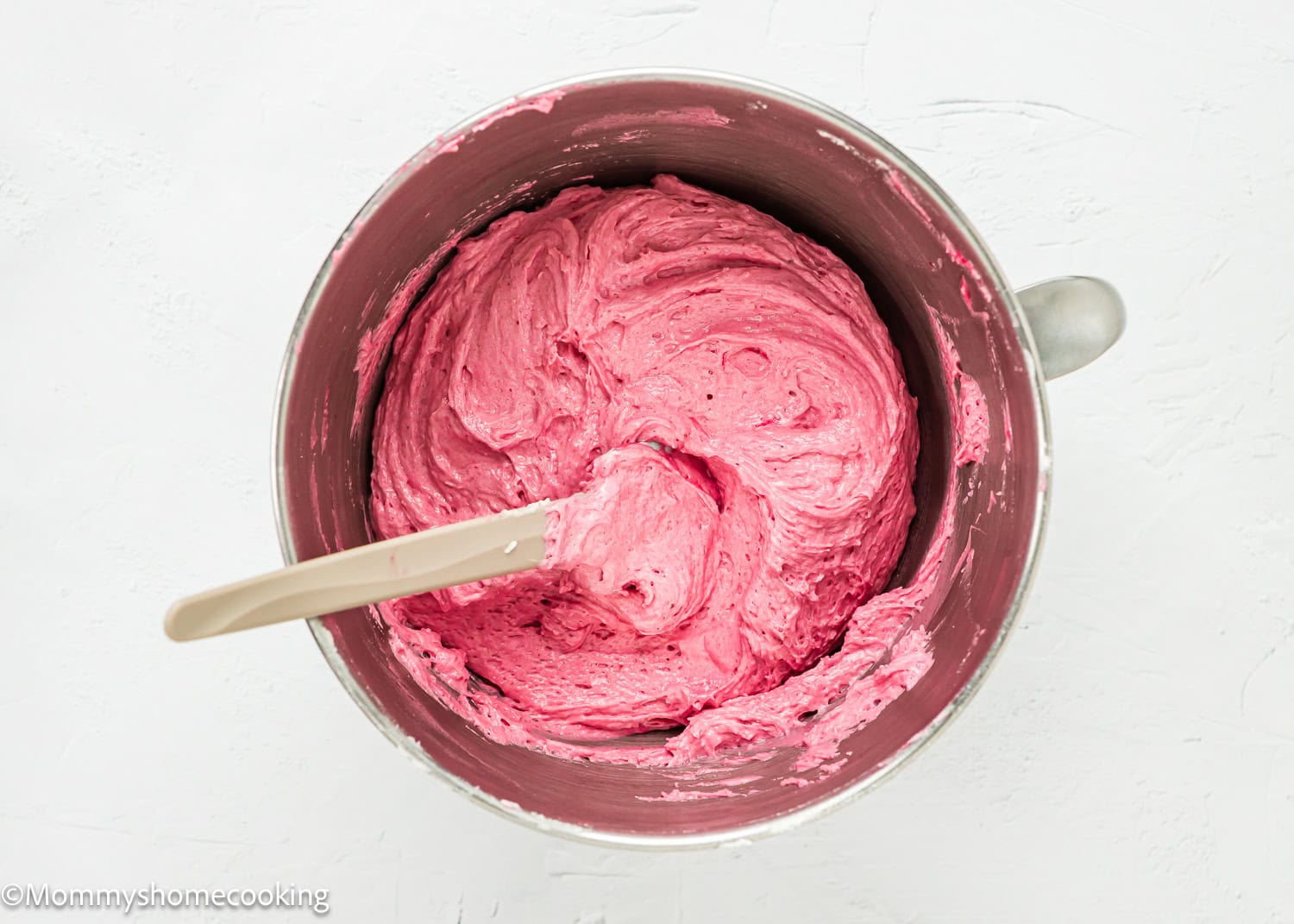 A mixing bowl filled with thick, bright pink frosting for Eggless Strawberry Crunch Cake, with a spatula resting inside.