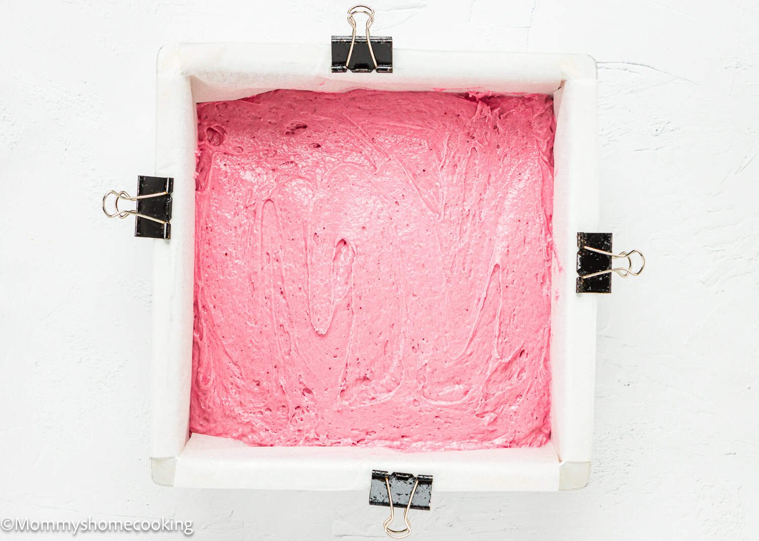 Square baking pan lined with parchment paper, secured with binder clips, filled with smooth pink batter for Eggless Strawberry Crunch Cake on a white surface.
