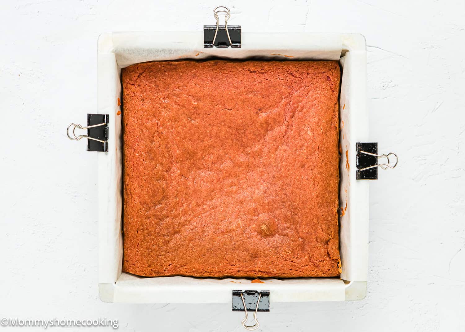 An Eggless Strawberry Crunch Cake in a parchment-lined baking pan secured with metal binder clips, viewed from above on a white background.