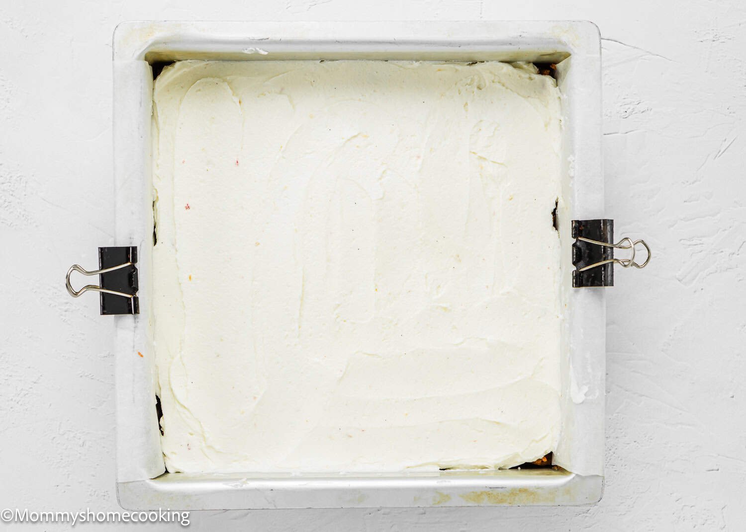 A square baking pan lined with parchment paper and clips, filled with a smooth layer of white frosting for an Eggless Strawberry Crunch Cake.